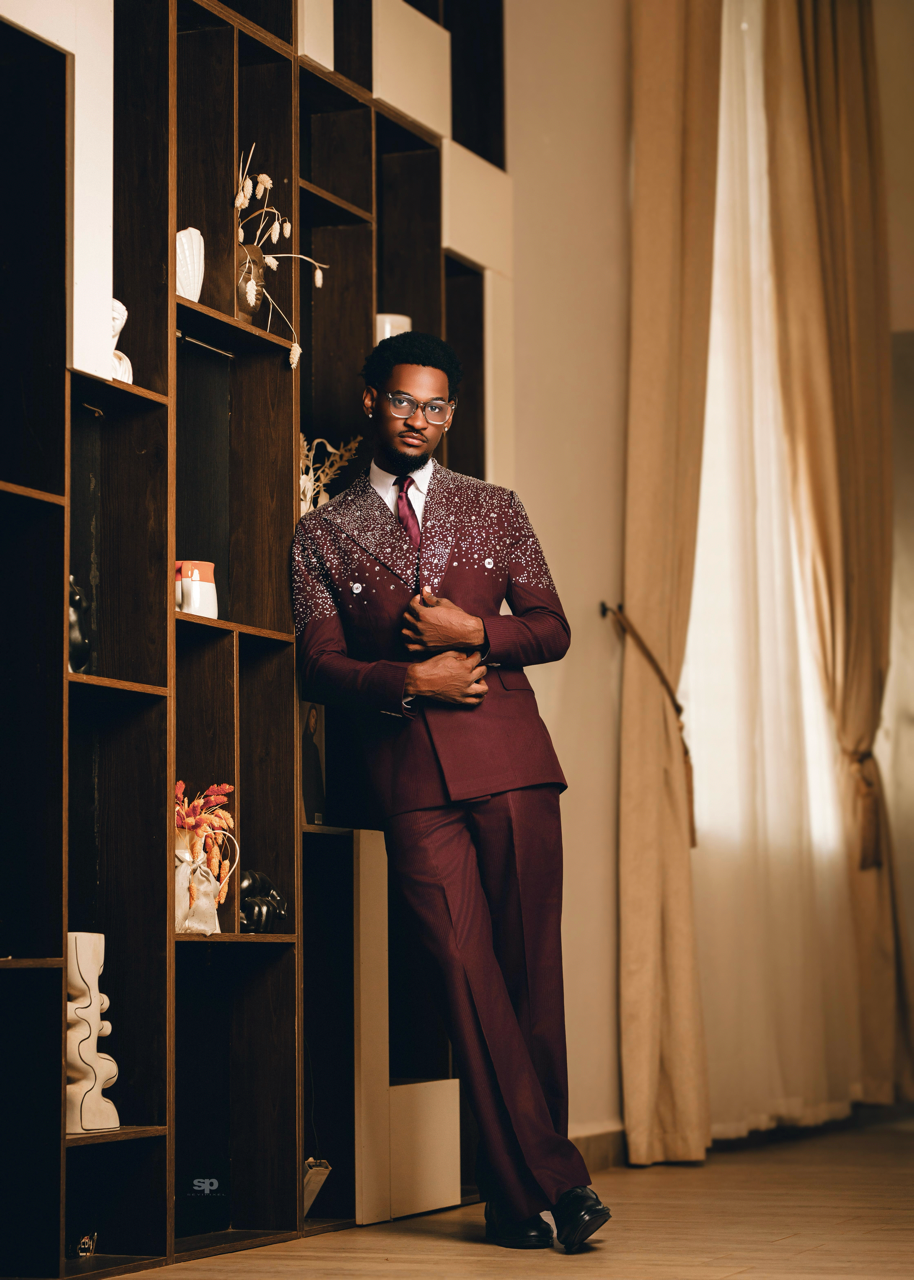 Man in a burgundy suit standing in a room with a bookshelf and curtains.
